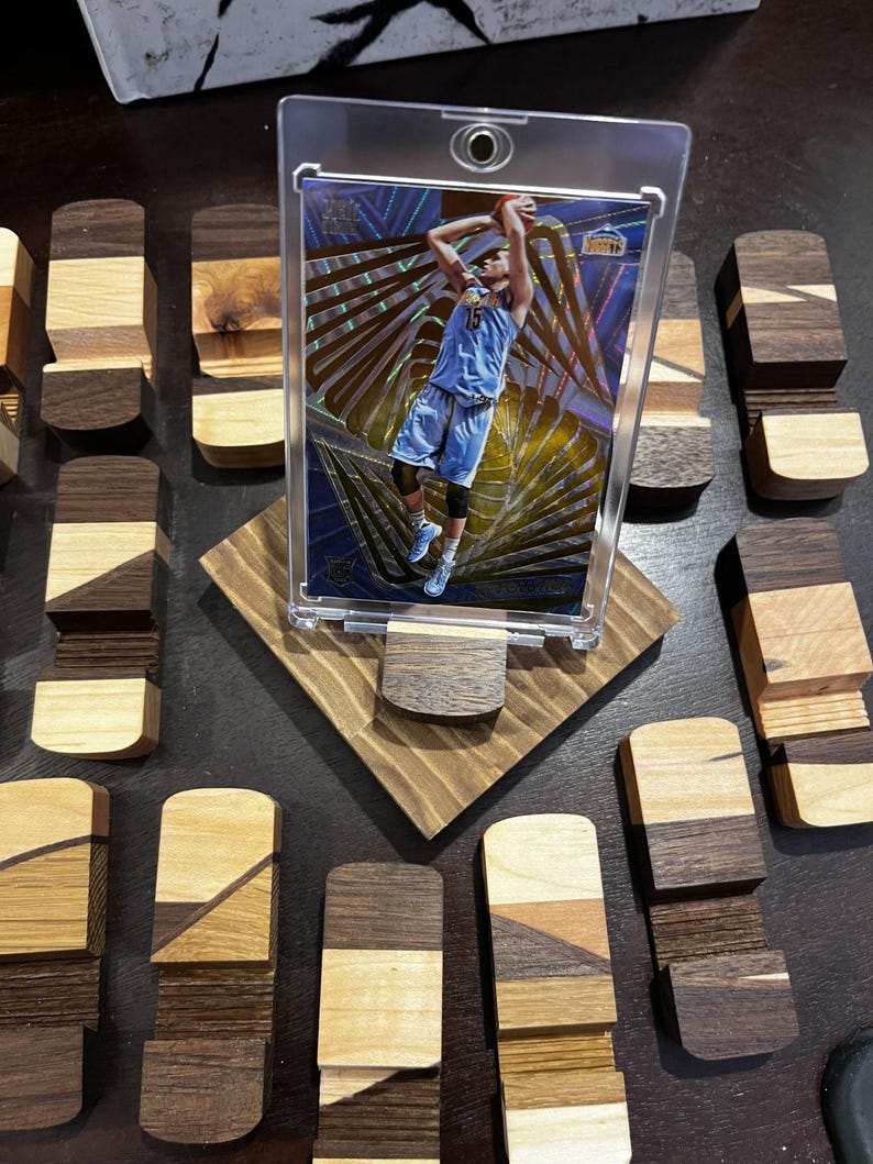 Puede incluir: Una tarjeta de baloncesto en una funda protectora transparente, exhibida en un soporte de madera. El soporte est&aacute; hecho de varios tonos de madera y est&aacute; rodeado de peque&ntilde;os bloques de madera. La tarjeta presenta a un jugador con uniforme azul.