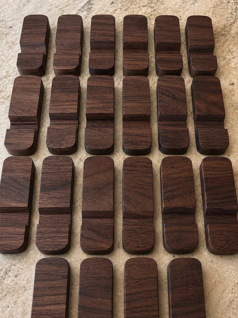 Puede incluir: Una colecci&oacute;n de veinti&uacute;n soportes de tel&eacute;fono de madera marr&oacute;n oscuro. Cada soporte es rectangular con una base curva y una ranura para sostener un tel&eacute;fono. La veta de la madera es visible en cada soporte.
