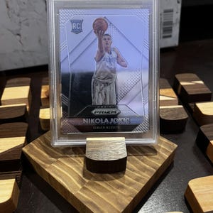 Puede incluir: Una tarjeta de baloncesto Panini Prizm Nikola Jokic de 2015, calificada como Mint 9, en una funda protectora. La tarjeta se muestra en un soporte de madera. La tarjeta muestra a Nikola Jokic jugando al baloncesto.