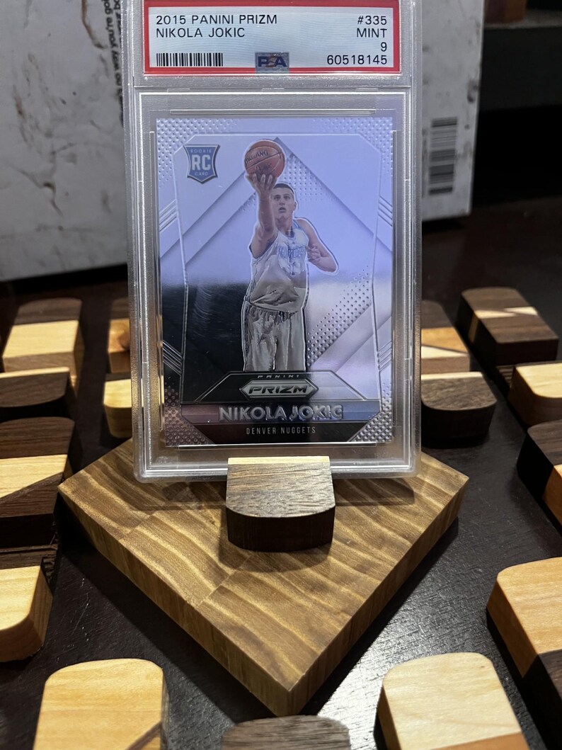 Puede incluir: Una tarjeta de baloncesto Panini Prizm Nikola Jokic de 2015, calificada como Mint 9, en una funda protectora. La tarjeta se muestra en un soporte de madera. La tarjeta muestra a Nikola Jokic jugando al baloncesto.