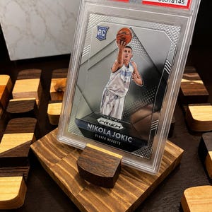 Puede incluir: Una tarjeta de novato de Nikola Jokic de 2015 Panini Prizm, calificada como Mint 9 por PSA, exhibida en un soporte de madera personalizado. La tarjeta presenta a Jokic con el uniforme de los Nuggets de Denver, lanzando un bal&oacute;n de baloncesto. El soporte es de madera clara y oscura.