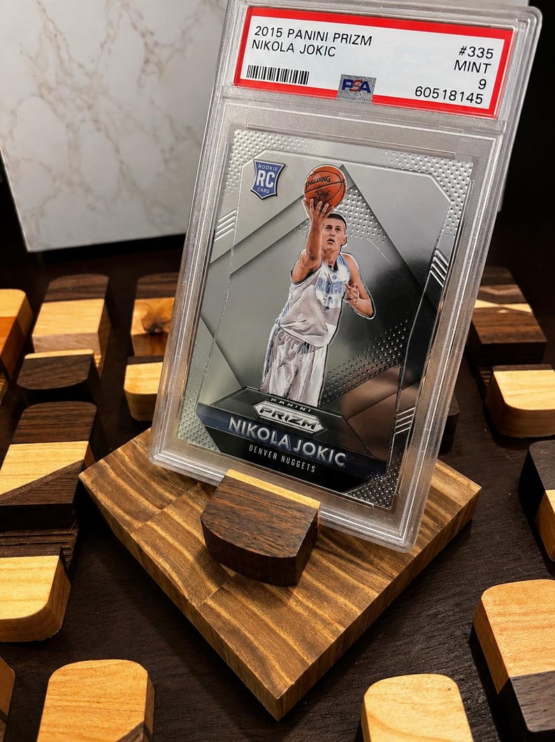 Puede incluir: Una tarjeta de novato de Nikola Jokic de 2015 Panini Prizm, calificada como Mint 9 por PSA, exhibida en un soporte de madera personalizado. La tarjeta presenta a Jokic con el uniforme de los Nuggets de Denver, lanzando un bal&oacute;n de baloncesto. El soporte es de madera clara y oscura.