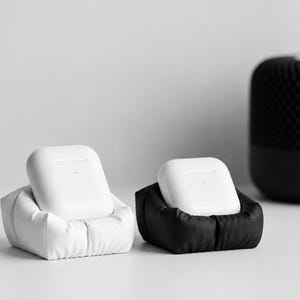 May include: Two white wireless earbud charging cases, each resting in a small, square, cushioned holder. One holder is white and the other is black, both featuring a soft, gathered fabric texture that cradles the rectangular charging cases securely.