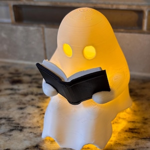 May include: A 3D-printed white ghost figurine with a smooth, layered texture, illuminated from within by a warm yellow light. The ghost has two oval-shaped eye cutouts and is holding an open, black book with visible horizontal line details.