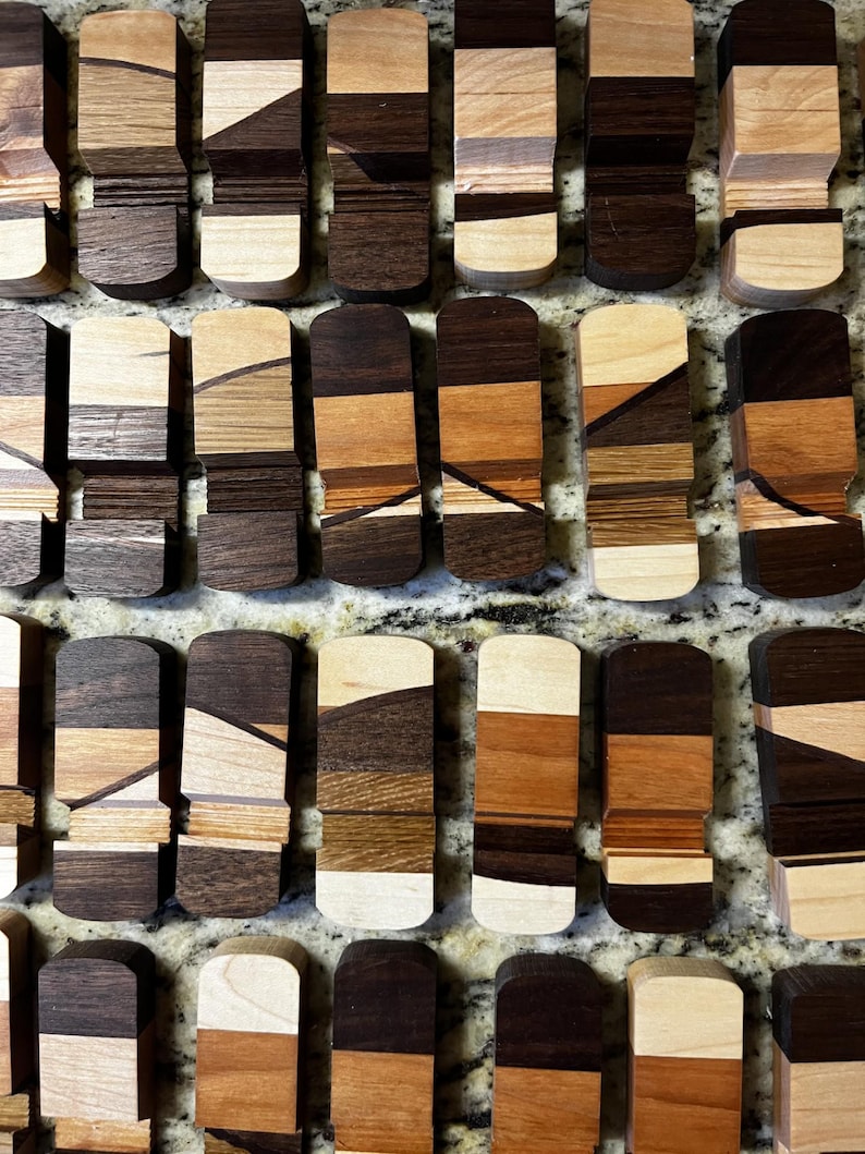 Puede incluir: Una colecci&oacute;n de piezas rectangulares de madera con extremos redondeados, cada una hecha de varios tipos de madera. Las piezas muestran una mezcla de tonos de madera claros y oscuros, incluyendo tonos de marr&oacute;n, beige y crema, dispuestos en un patr&oacute;n de mosaico. Las piezas est&aacute;n dispuestas sobre una superficie moteada.