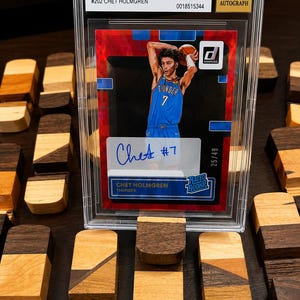 Puede incluir: Una tarjeta de baloncesto de Chet Holmgren, calificada por Beckett, con un dise&ntilde;o rojo y azul. La tarjeta es una firma Donruss Rated Rookies 2022-23, Holo Red Laser, con el texto "Chet #7" y "25/49". La tarjeta est&aacute; calificada con 9 Mint y 10 Autograph.