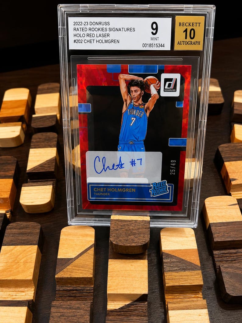 Puede incluir: Una tarjeta de baloncesto de Chet Holmgren, calificada por Beckett, con un dise&ntilde;o rojo y azul. La tarjeta es una firma Donruss Rated Rookies 2022-23, Holo Red Laser, con el texto "Chet #7" y "25/49". La tarjeta est&aacute; calificada con 9 Mint y 10 Autograph.