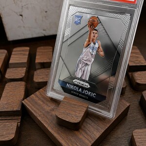 Puede incluir: Una tarjeta de baloncesto Nikola Jokic 2015 Panini Prizm, calificada como Mint 9, en un protector. La tarjeta muestra a Jokic jugando al baloncesto. La tarjeta se exhibe en un soporte de madera.