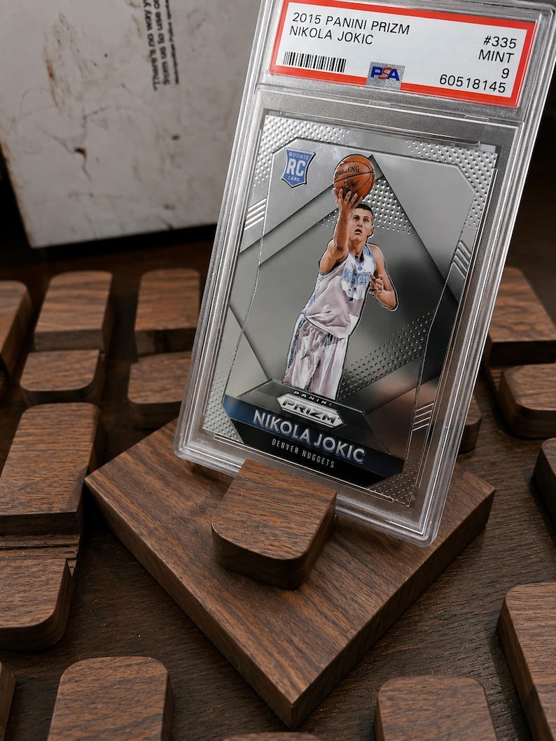 Puede incluir: Una tarjeta de baloncesto Nikola Jokic 2015 Panini Prizm, calificada como Mint 9, en un protector. La tarjeta muestra a Jokic jugando al baloncesto. La tarjeta se exhibe en un soporte de madera.