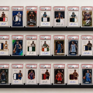 May include: A collection of twenty-four professional basketball trading cards, each encased in a clear protective PSA slab, displayed on three horizontal black wall-mounted shelves. The cards feature various player portraits, team jersey patches, and signatures.
