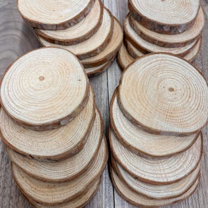 Könnte beinhalten: Mehrere Stapel runder Untersetzer aus Naturholz. Jeder Untersetzer hat eine hellbraune Mitte mit dunkleren braunen Rändern, die die Jahresringe des Baumes zeigen. Die Untersetzer sind auf einer Holzoberfläche angeordnet und bieten eine rustikale und organische Ästhetik.