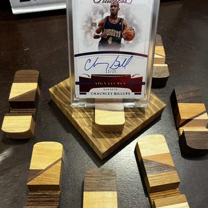 Puede incluir: Una tarjeta de baloncesto de Chauncey Billups en una funda protectora, exhibida en un soporte de madera. La tarjeta es una tarjeta de firma "Flawless", numerada 12/15. Varios bloques de madera decorativos peque&ntilde;os con diferentes patrones de vetas de madera rodean el soporte.
