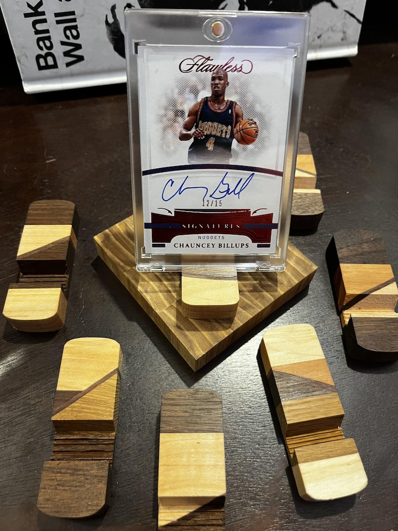 Puede incluir: Una tarjeta de baloncesto de Chauncey Billups en una funda protectora, exhibida en un soporte de madera. La tarjeta es una tarjeta de firma "Flawless", numerada 12/15. Varios bloques de madera decorativos peque&ntilde;os con diferentes patrones de vetas de madera rodean el soporte.