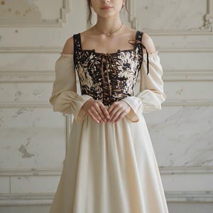 May include: A cream-colored dress with long sleeves and a floral patterned bodice. The bodice is brown and black with a lace-up front. The dress has off-the-shoulder straps and a flowing skirt, creating a vintage aesthetic.