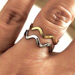 Peut inclure: Deux bagues en forme de vagues, une dorée et une argentée, sont superposées sur un doigt. Les bagues ont un design simple et minimaliste. L'arrière-plan est flou, attirant l'attention sur les bijoux.