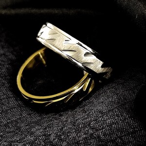 Könnte beinhalten: Zwei Ringe, einer goldfarben und einer silberfarben, werden vor einem dunklen Hintergrund gezeigt. Der goldfarbene Ring hat ein geometrisches Muster, während der silberfarbene Ring ein strukturiertes Design aufweist. Die Ringe sind wahrscheinlich für eine Hochzeit oder Verlobung.