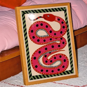 Könnte beinhalten: Ein gerahmter Kunstdruck mit einer stilisierten rosa Schlange mit schwarzen Punkten und einem roten Kopf. Das Kunstwerk ist vor einem cremefarbenen Hintergrund platziert und mit einem Holzrahmen versehen. Der Druck wird auf einem weißen Teppich in der Nähe eines rosa Bettes ausgestellt.