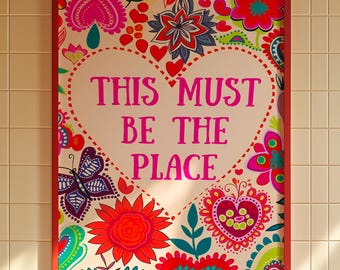 May include: A framed art print with a red border. The artwork features a heart-shaped design with the text "THIS MUST BE THE PLACE" in pink. The background is filled with colorful floral and butterfly patterns in shades of red, pink, and green.