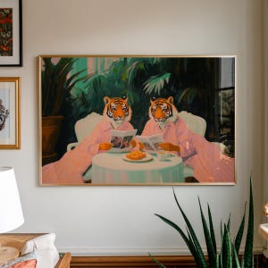 May include: A framed art print depicts two tigers in pink robes seated at a table, reading. The tigers have orange and black striped faces. A small table holds pastries and a cup of coffee. The background features lush green foliage.