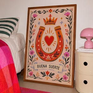 Puede incluir: Una lámina rectangular enmarcada que presenta una herradura naranja central y un corazón rojo, rematados con una corona naranja. La obra incluye bordes florales, pequeñas estrellas y una cinta en la parte inferior con el texto BUENA SUERTE.