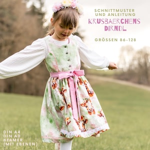 May include: A girl in a traditional dirndl dress, featuring a green bodice, white blouse, and a skirt with a cow print. The dress is accented with a pink sash and floral crown. Text on the image reads "KRUSBAERCHENS DIRNDL" and "GRÖSSEN 86-128".