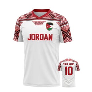 May include: White soccer jersey with red accents and the word "JORDAN" in red block letters. The jersey features a crest with the Jordanian flag and a patterned design on the shoulders and sleeves. A smaller jersey shows "YOUR NAME" and the number 10.