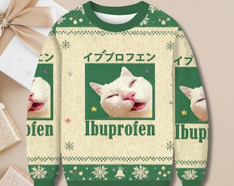 May include: A cream-colored Christmas sweater with a green collar and cuffs. The sweater features a white cat face graphic with the word "Ibuprofen" below it. The Japanese characters for "Ibuprofen" are above the cat graphic. The sweater has a festive snowflake and star pattern.