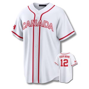 Pode incluir: Camisola de baseball branca com detalhes vermelhos e a palavra "CANADA" no peito. A camisola tem uma frente abotoada, mangas curtas e um pequeno emblema da bandeira canadiana. Uma camisola menor com "YOUR NAME" e o número 12 também é mostrada.