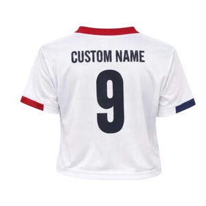 May include: White athletic jersey with red and navy blue trim. The back of the jersey features the number 9 in navy blue and the text "CUSTOM NAME" above it. The jersey has a round neck and short sleeves.