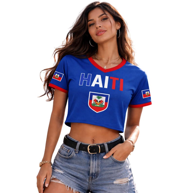 May include: A blue crop top with red trim and the word "HAITI" in white and red. The shirt features the Haitian flag and a crest. The model is wearing denim shorts and a black belt.