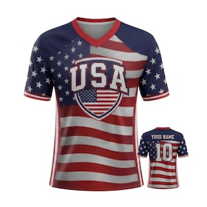 May include: A red, white, and blue USA soccer jersey with a V-neck design. The front features the word "USA" above a shield with the American flag. The sleeves and shoulders have a blue background with white stars. A smaller jersey is shown on the right.