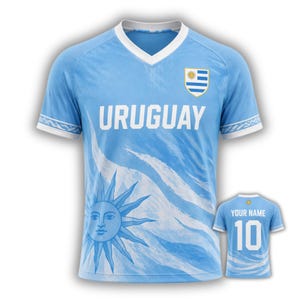 Peut inclure: Maillot de football de l'Uruguay bleu clair avec un col en V et des manches blanches. Le maillot présente le mot "URUGUAY" en blanc, un motif de soleil et l'emblème du drapeau uruguayen. Un maillot plus petit affiche "YOUR NAME" et le numéro 10.