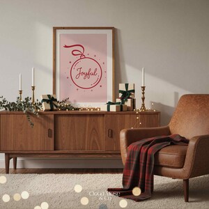 May include: A wooden cabinet with a framed print that says "Joyful" in red script. The print is a pink background with a red Christmas ornament design. Several wrapped gifts with green ribbons and candles are on the cabinet. A brown leather armchair and a red plaid blanket are in the scene.