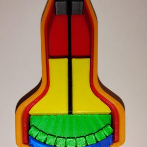 May include: A multi-colored 3D printed object shaped like a bell or rocket, featuring an orange outer shell, a red inner layer, two yellow central blocks, a green row of small square segments, a blue base, and a thin black vertical rod.