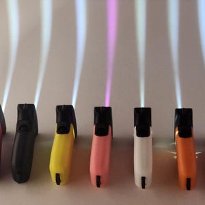 May include: Seven handheld flashlights in a row, each featuring a different colored casing including pink, black, yellow, light pink, white, orange, and purple. Each flashlight emits a vertical beam of light upward, with one beam appearing pink.