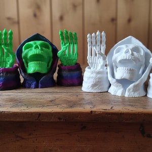 Könnte beinhalten: Eine Sammlung von 3D-gedruckten Schädel-Figuren. Zwei grüne Schädel mit Kapuzen und Händen und zwei weiße Schädel mit Kapuzen und Händen. Die Hände sind in einer Grußgeste. Die Figuren stehen auf einer Holzoberfläche.