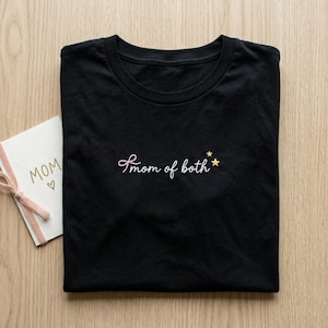 May include: Black t-shirt with the embroidered phrase "mom of both" in a cursive font, accompanied by two small gold stars. A card with the word "MOM" and a heart is visible.