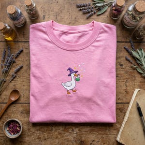 May include: Pink t-shirt featuring an embroidered cartoon goose wearing a purple witch hat and carrying a green potion bottle. The goose is walking with a magical trail. The shirt is folded on a wooden surface.