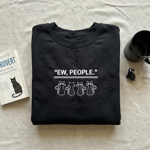 May include: Black sweatshirt with the phrase "EW, PEOPLE." in white text above a row of four embroidered cat silhouettes. A black mug, a small black cat figurine, and a book with a cat illustration are also visible.