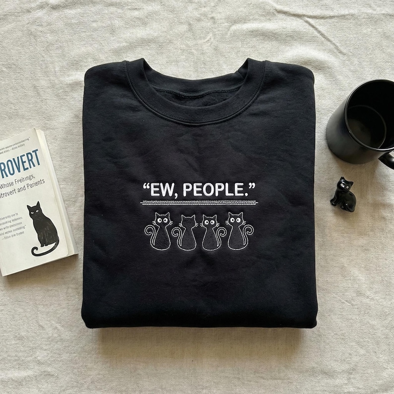 May include: Black sweatshirt with the phrase "EW, PEOPLE." in white text above a row of four embroidered cat silhouettes. A black mug, a small black cat figurine, and a book with a cat illustration are also visible.