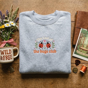 May include: A pale grey sweatshirt featuring the embroidered phrase "The Last of the bugs club" and two ladybugs. The sweatshirt is displayed with flowers, a mug with "WILD & FREE", and a postcard.