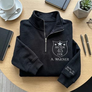 May include: Black quarter-zip sweatshirt with a silver zipper and embroidered white design. The design includes the text "SECTOR 45 CCR" and "A. WARNER" within a shield. The sleeve has the text "Ignite, my love. Ignite."
