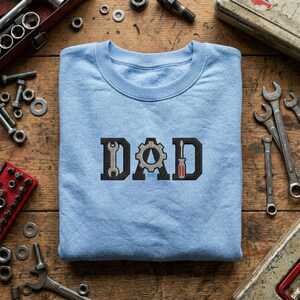 May include: A pale blue crew neck sweatshirt displaying the word "DAD" in a black and grey design. The letters are formed with tools: a wrench, a gear, and a screwdriver. The sweatshirt is on a wooden surface with various tools.