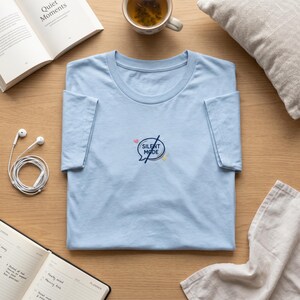 May include: A light blue t-shirt with the words "SILENT MODE" in a circular design. The design includes a heart, a lightning bolt, and a star. The shirt is folded neatly on a wooden surface, with a book, tea cup, and headphones nearby.
