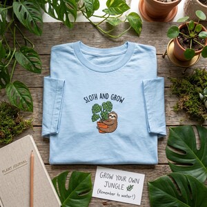May include: A pale blue t-shirt featuring the text "SLOTH AND GROW" and an image of a sloth with a plant. A card reads "GROW YOUR OWN JUNGLE (Remember to water!)". The shirt is displayed with plants and a journal on a wooden surface.
