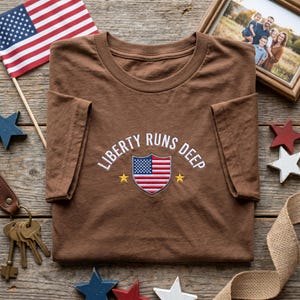 Könnte beinhalten: Ein braunes T-Shirt mit dem gestickten Schriftzug "LIBERTY RUNS DEEP" und einem amerikanischen Flaggenemblem. Das Shirt liegt auf einer Holzoberfläche, mit einer amerikanischen Flagge und dekorativen Sternen.