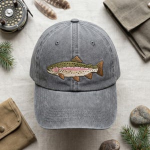 May include: Gray baseball cap with an embroidered rainbow trout design. The cap is made of a soft, washed fabric and features a curved brim. The trout is detailed with shades of green, pink, and brown, perfect for fishing enthusiasts.