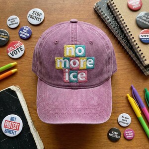 May include: A faded pink baseball cap featuring the embroidered phrase "no more ice" in a block letter design. The image includes protest badges and pens on a wooden surface.