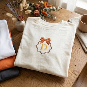 May include: Off-white t-shirt with an embroidered design of a bow and the letter "D" in yellow. The design is framed by a brown border. Several folded t-shirts in white, orange, and grey are stacked to the left.