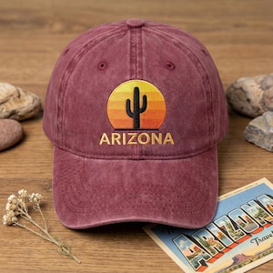May include: A maroon baseball cap with a curved peak. The cap has an embroidered design of a black cactus silhouette against a yellow and orange sunset, with the word "ARIZONA" embroidered below in gold.